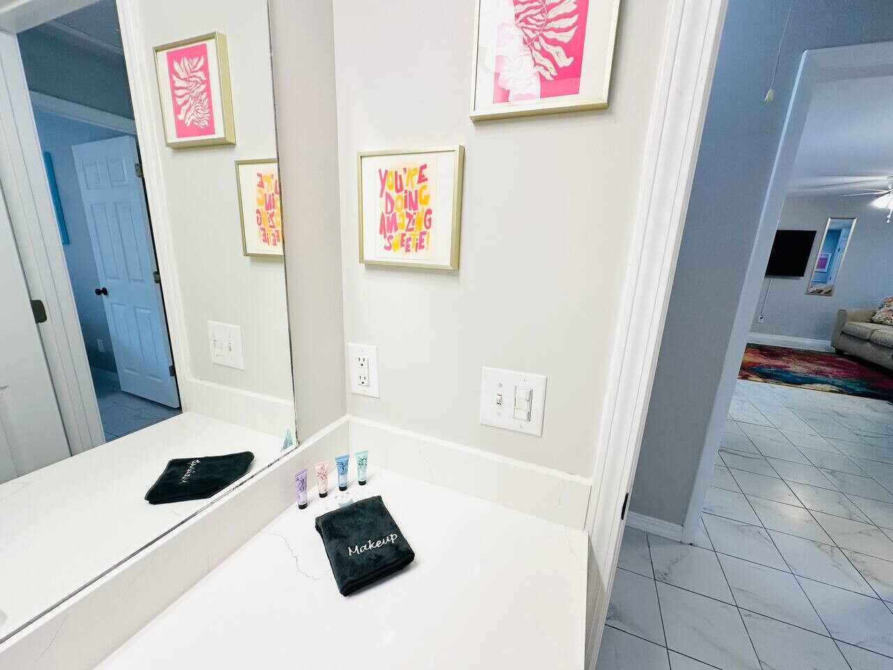 A bathroom with a large mirror, a vanity with makeup items, and framed artwork on the wall.