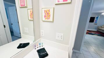 A bathroom with a large mirror, a vanity with makeup items, and framed artwork on the wall.