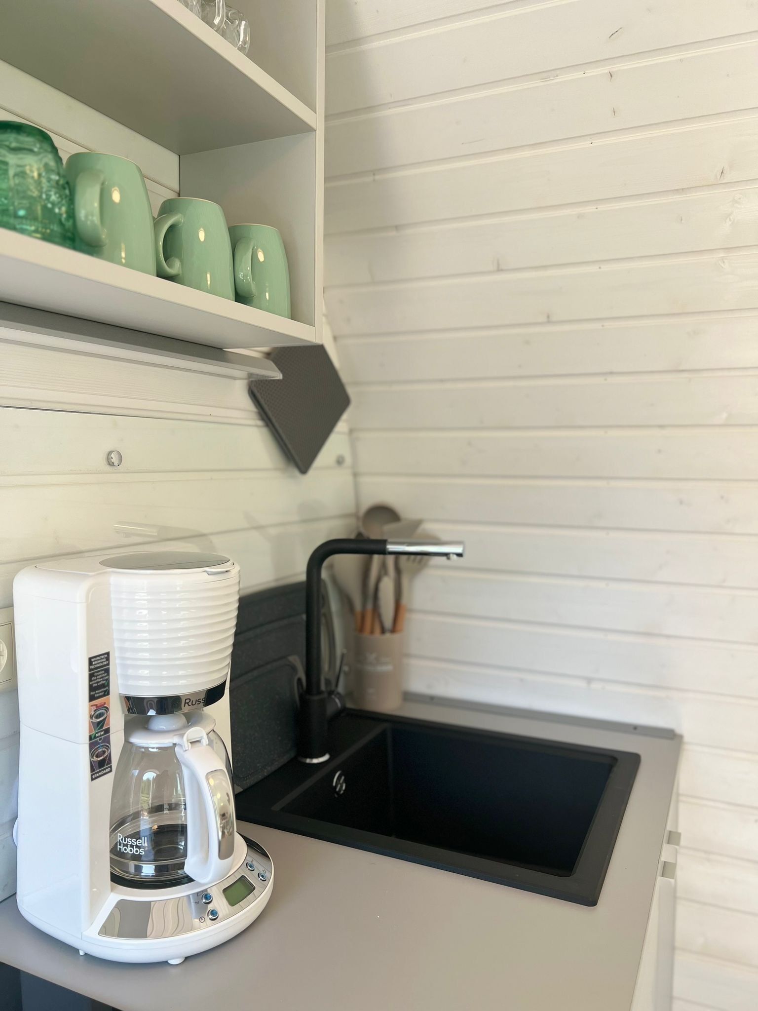 A kitchen with a coffee maker, a sink, and green cups on a shelf.