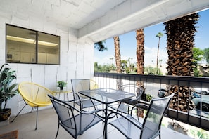 A balcony with a table and chairs, a potted plant, and a view of palm trees and a car. - Palm Canyon Retreat, 2bd 2ba with Pool and Patio (Palm Springs)