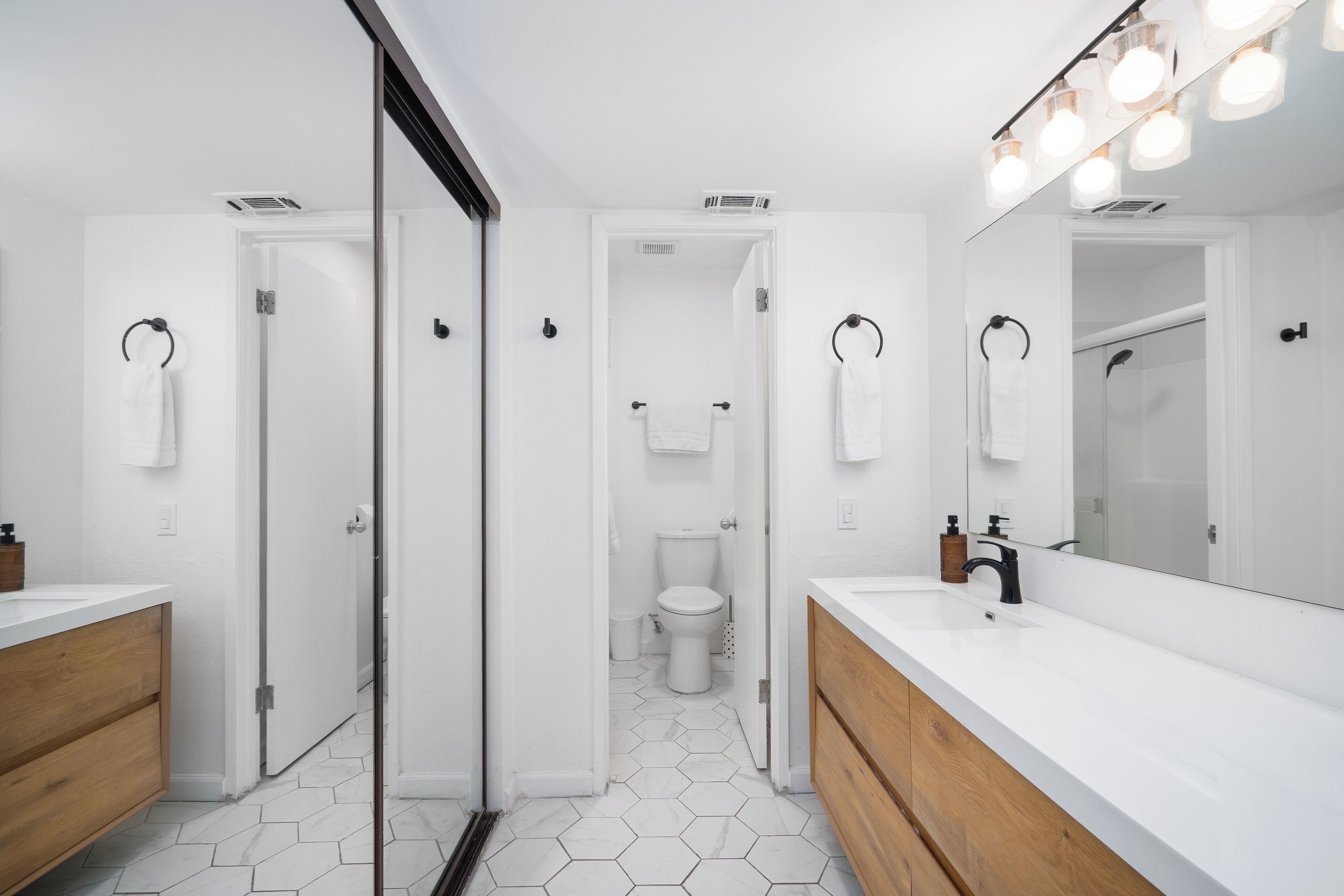 A modern bathroom with white walls, hexagonal tile flooring, a large mirror with lights, and two sinks.