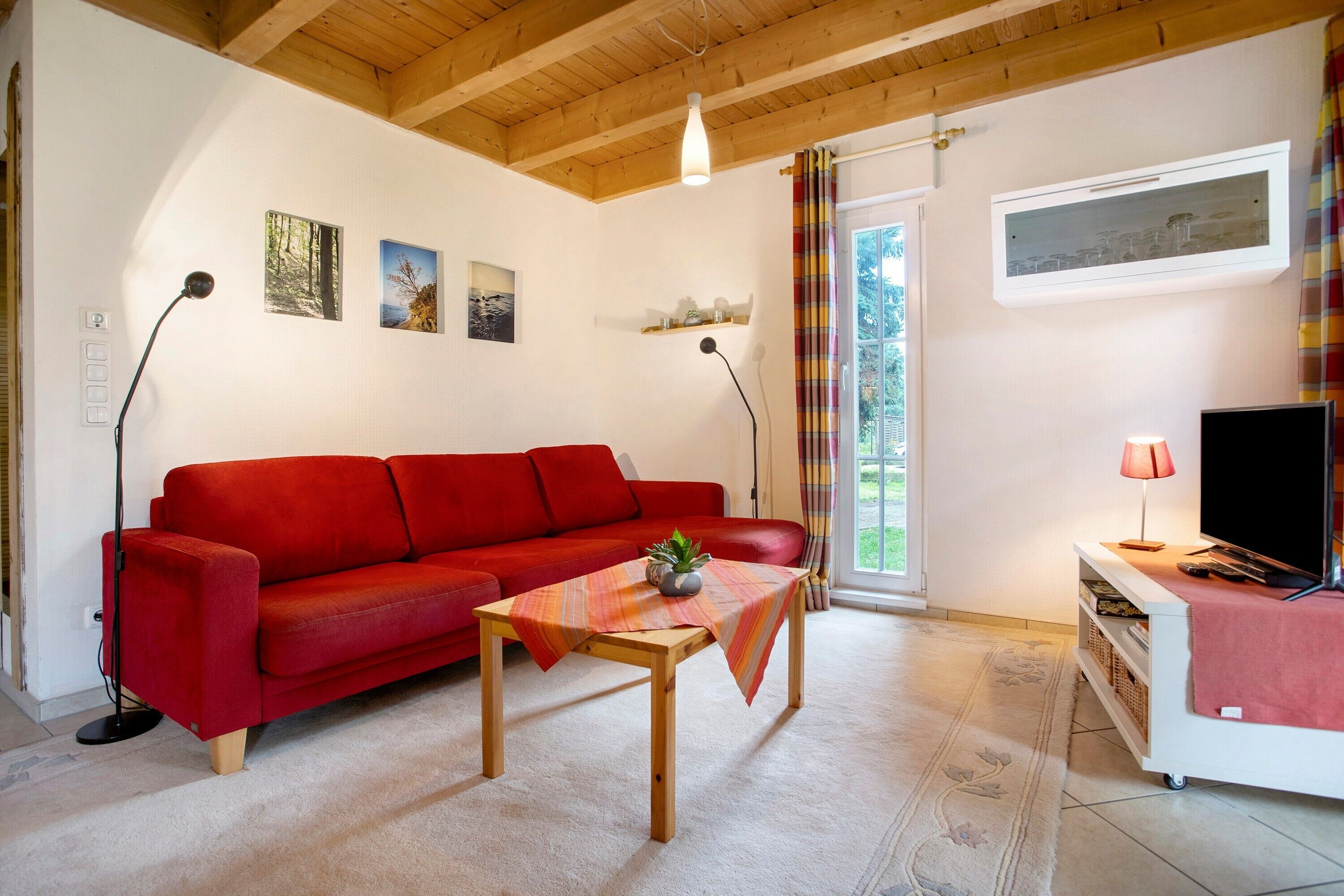 Un canapé rouge, une table en bois avec une plante, un téléviseur posé sur un meuble blanc et une fenêtre avec des rideaux.
