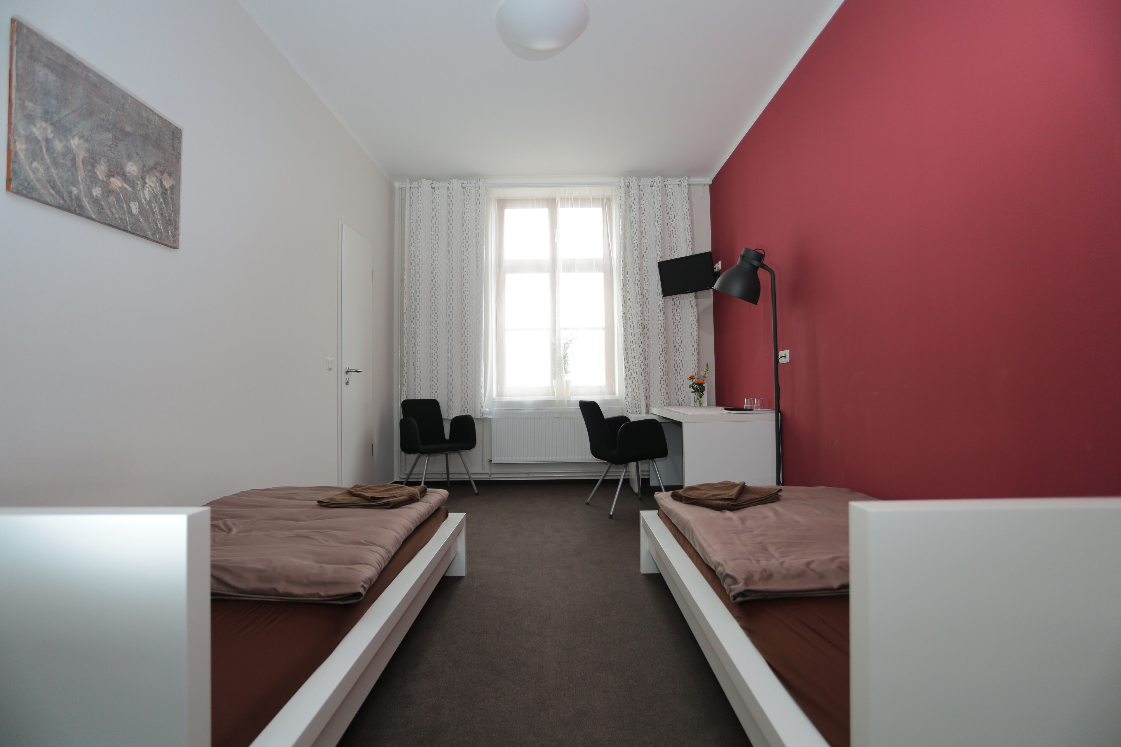 Ein Zimmer mit zwei Einzelbetten, einem Schreibtisch, zwei Stühlen und einem Fenster mit Vorhängen. Eine Wand ist rot gestrichen.