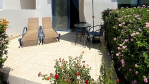 Ein weißes Gebäude mit einer überdachten Terrasse, auf der sich ein Tisch und Stühle, ein blauer Sonnenschirm und ein kleiner Garten mit roten und pinkfarbenen Blumen befinden.