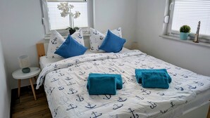 Un dormitorio con una cama que tiene una colcha con motivos náuticos, dos almohadas, una mesita de noche con una lámpara y una ventana con persianas.