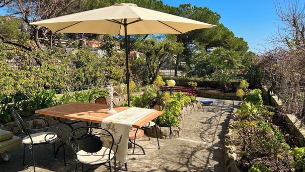 An outdoor dining area with a table and chairs, surrounded by lush greenery and a stone pathway. - Garden view apartment in Sanremo with veranda (Sanremo)