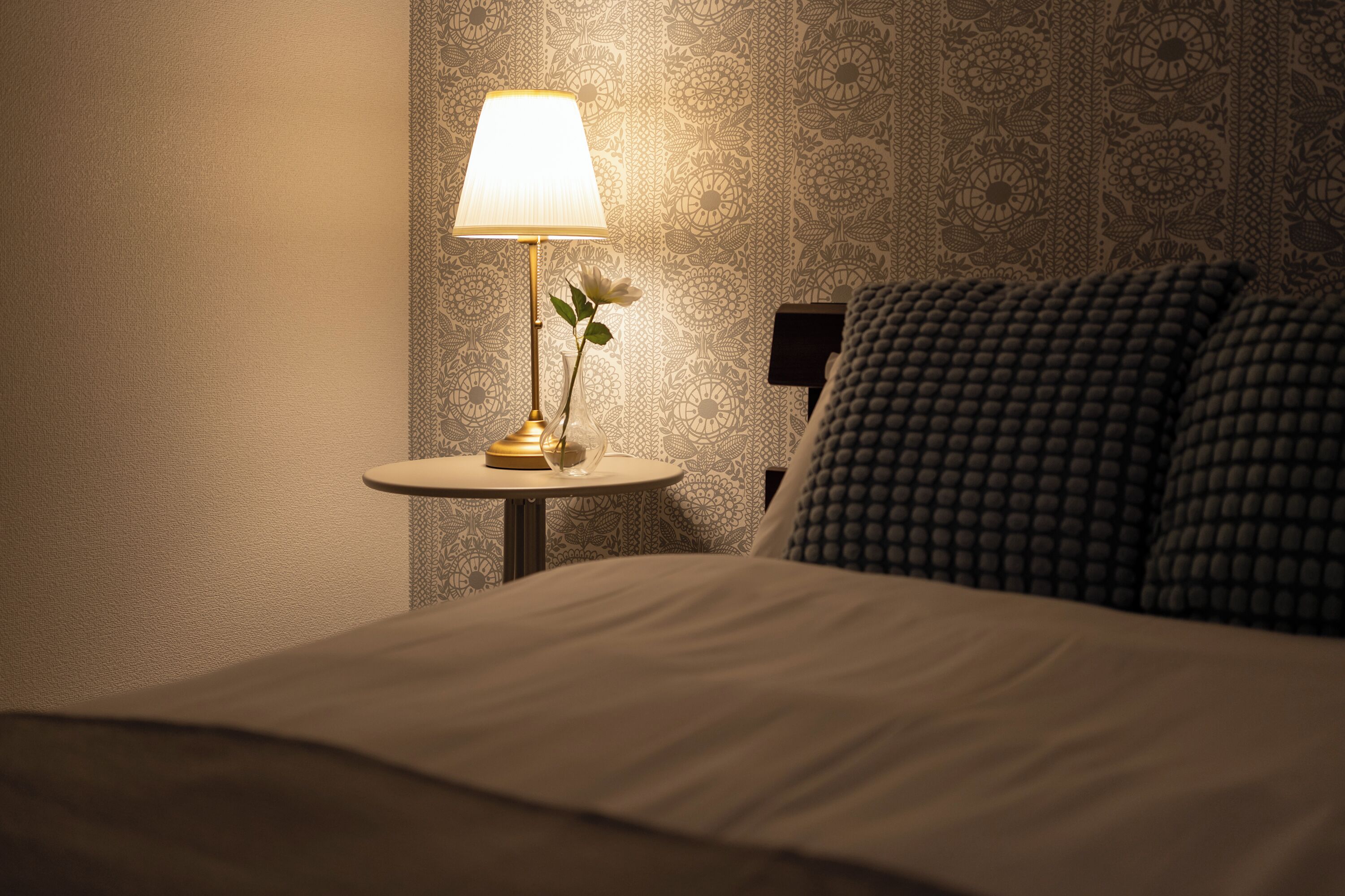A neatly made bed with a white comforter, a small round table with a lamp and a vase of flowers, and a patterned wallpaper on the wall.
