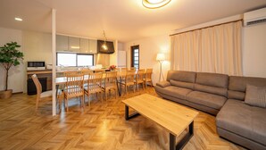 A modern living room with a wooden herringbone floor, a brown sofa, a wooden coffee table, and a dining area with wooden chairs and a table. - ALLURE BEPPU (Beppu)