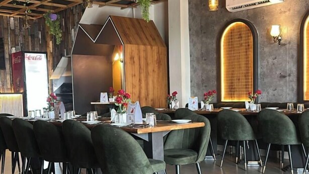 A restaurant with a modern interior, featuring a wooden ceiling with hanging plants, a central seating area with green upholstered chairs, and a bar area with a Coca-Cola sign. - The Divine Cloud Inn (Aligarh)