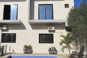 Um edifício moderno com janelas de dois andares, aparelhos de ar condicionado e uma pequena área de piscina com plantas em vasos.