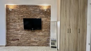 A living room with a stone wall, a flat-screen TV, a wardrobe, and tiled flooring. - 157 LIM - Stay Sunsea Studio (Limassol)