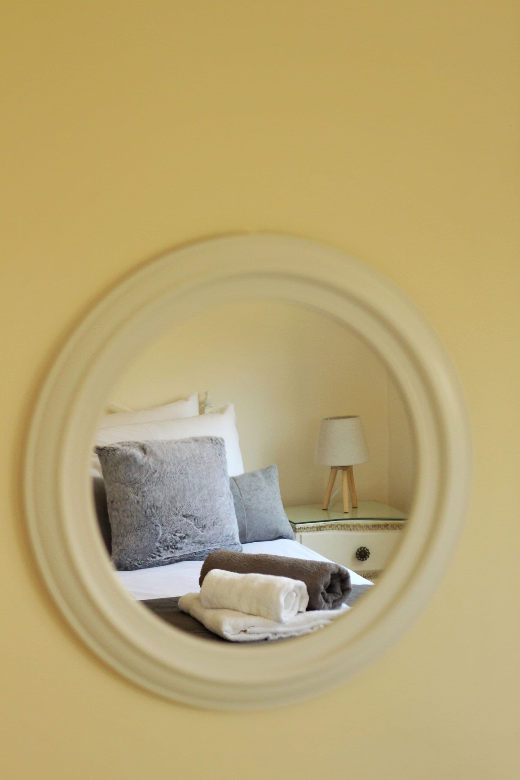 A circular mirror reflecting a bed with pillows and a nightstand with a lamp.