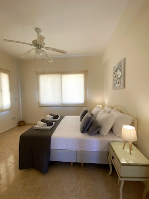A bedroom with a bed, a bedside table with a lamp, a ceiling fan, and a window with blinds.