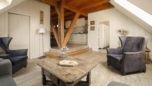 Ein Loft-Zimmer mit freiliegenden Holzbalken, einem Esstisch, zwei Sesseln und einer Treppe, die zu einer erhöhten Ebene führt.