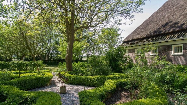 Ein Garten mit Kiesweg, ordentlich geschnittenen Hecken und einem Gebäude mit Strohdach.