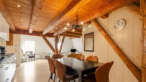 Een eethoek met een houten tafel en stoelen, een klok aan de muur en een keuken op de achtergrond.