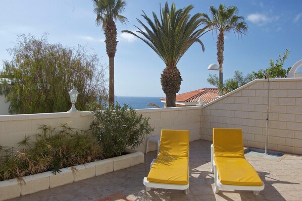 A rooftop terrace with two yellow sun loungers, a palm tree, and a view of the sea.
