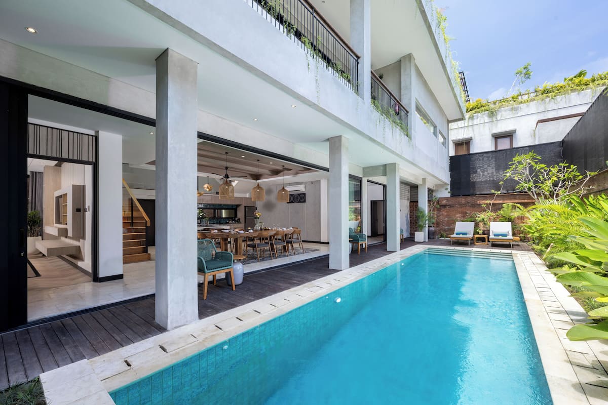 Un área de piscina moderna con vista despejada, sillones de descanso y un comedor con una escalera que lleva a un nivel superior.