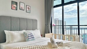 Une chambre d’hôtel avec un grand lit, une tête de lit grise et une vue sur la ville grâce à de grandes fenêtres.