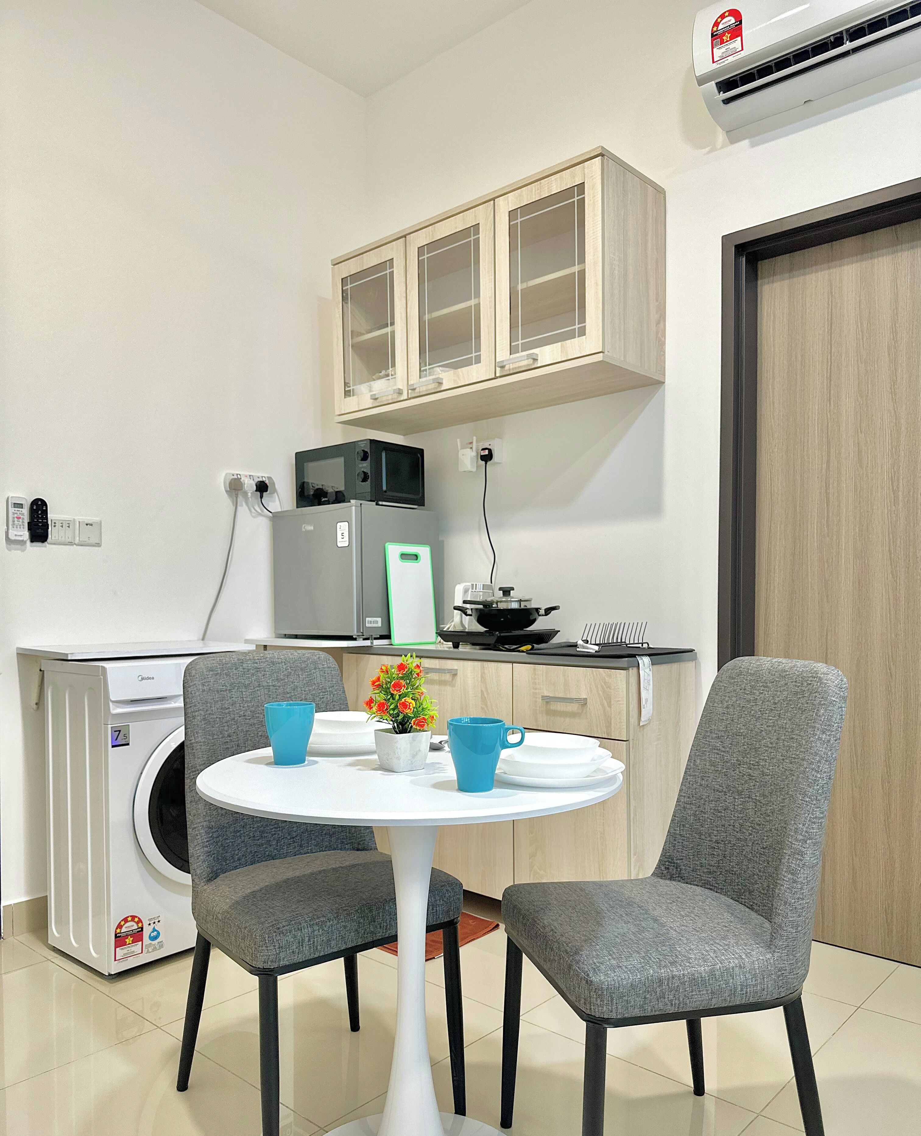 Een compacte keuken met een magnetron, een klein spoelgedeelte en een kastje boven het aanrecht. Er staat een eettafel met twee stoelen en een kleine potplant.