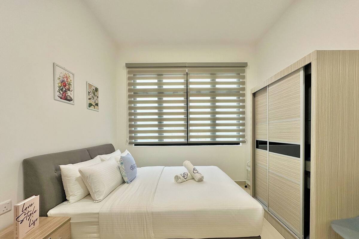 Un dormitorio ordenado con cama, almohadas, una mesita de noche, un armario y una ventana con persianas.