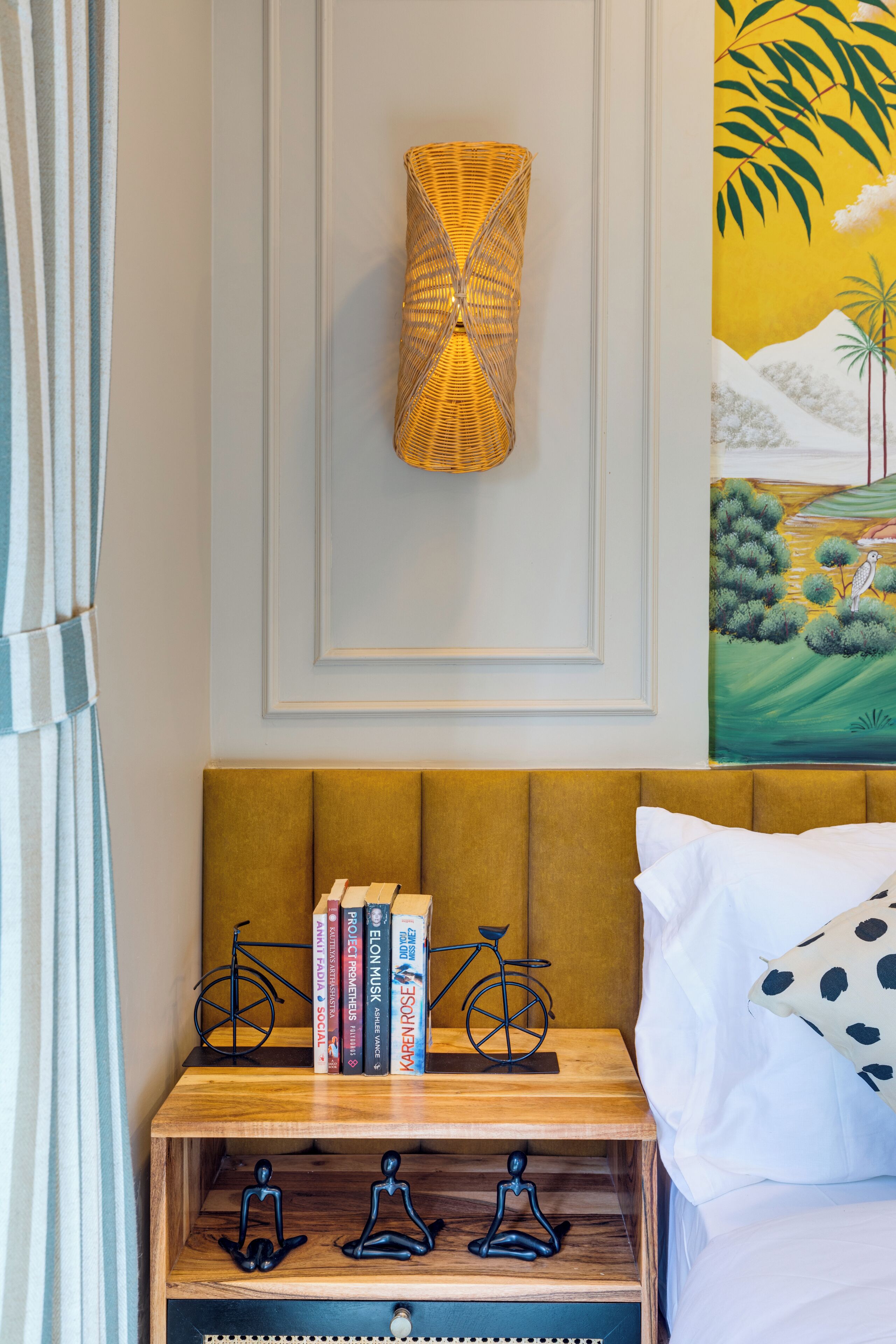 A neatly arranged bedroom with a wooden headboard, a bedside table with books and a bicycle, and a wall-mounted lamp.