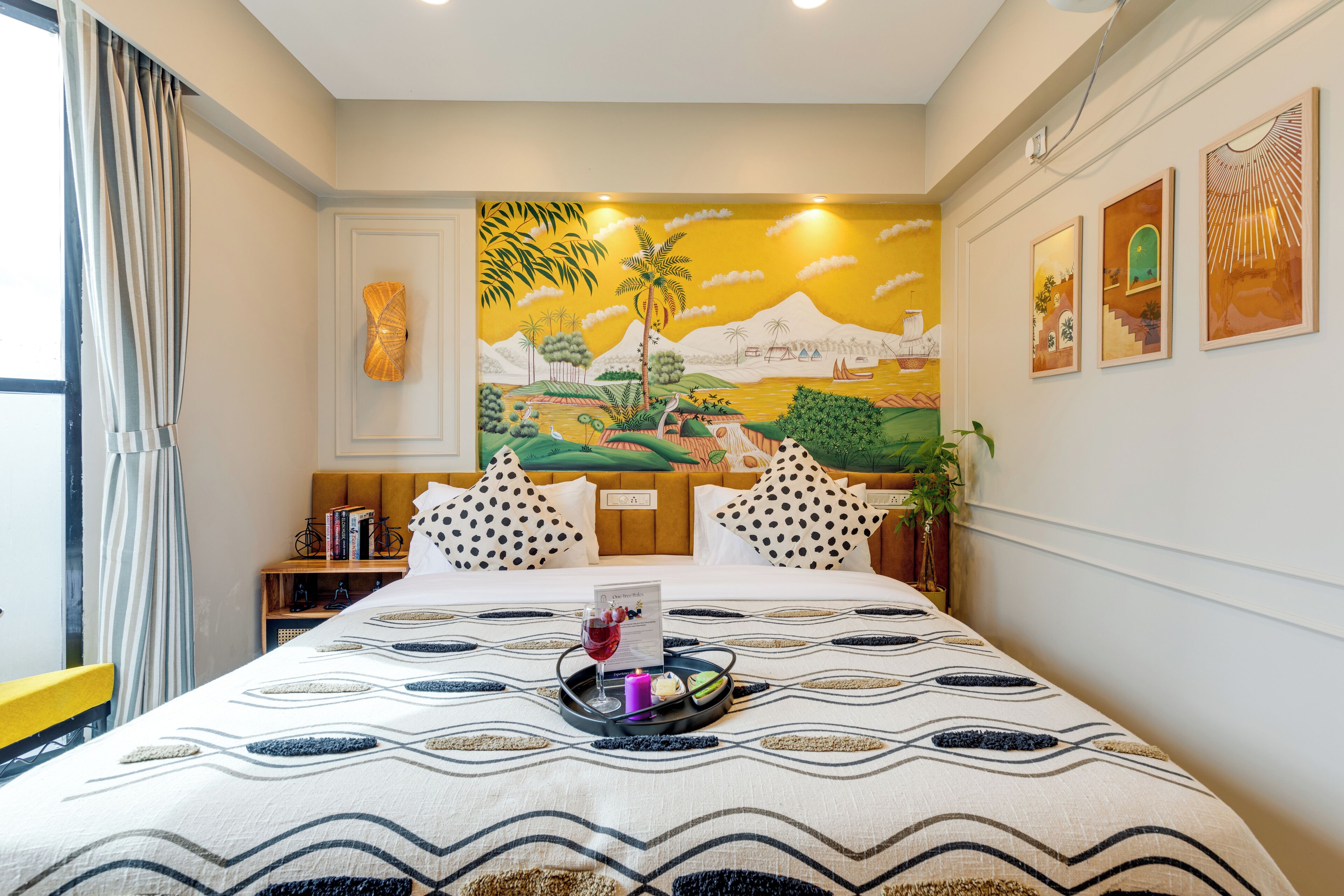 A hotel room with a bed, a nightstand with books, a wall mural, and a window with curtains.