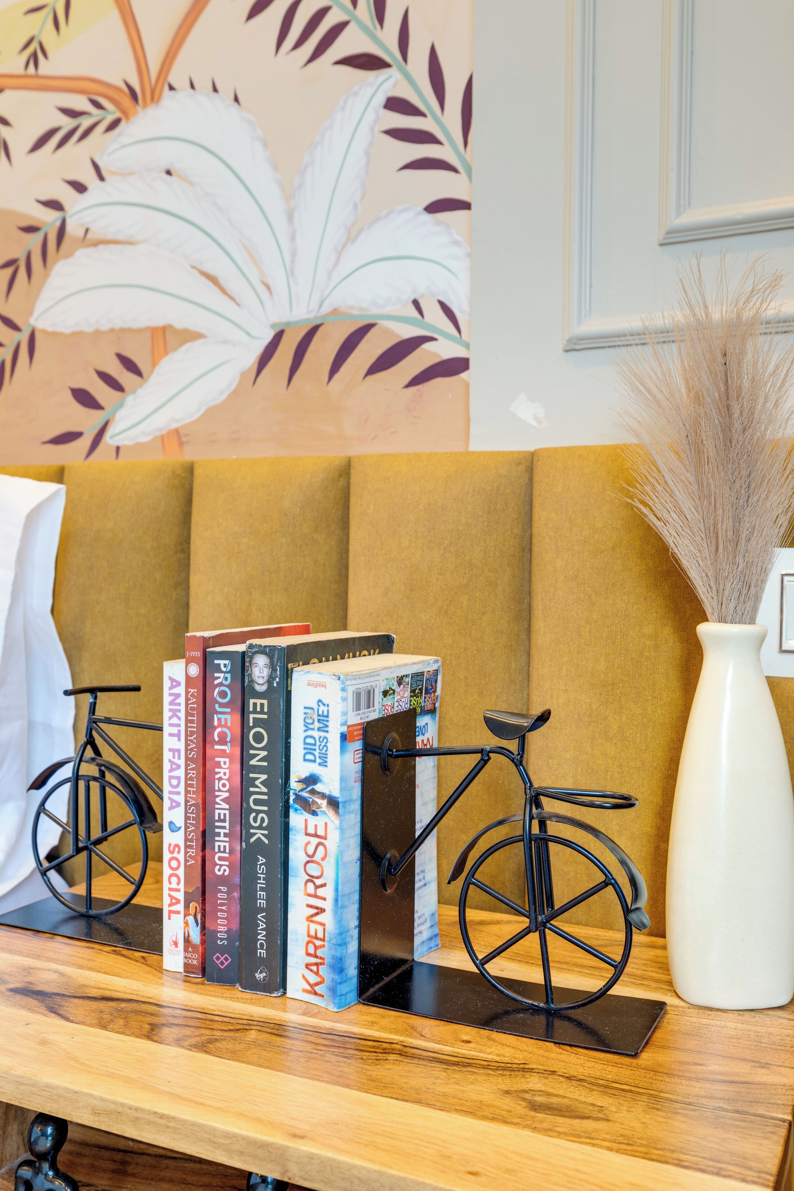 Ein Bücherregal mit Büchern und einem Fahrrad, eine weiße Vase mit Trockenpflanzen und eine Wand mit Blätermuster.