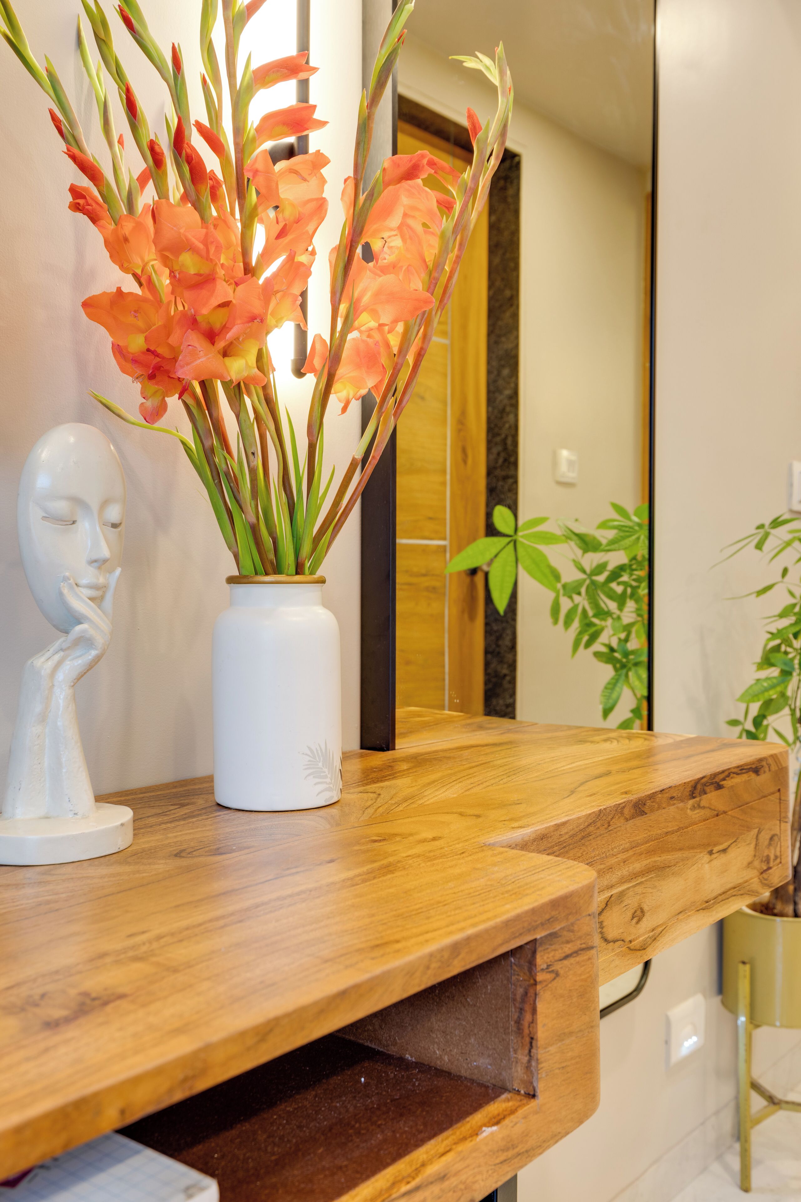 A wooden counter with a white vase containing orange flowers, a white bust sculpture, and a green plant.