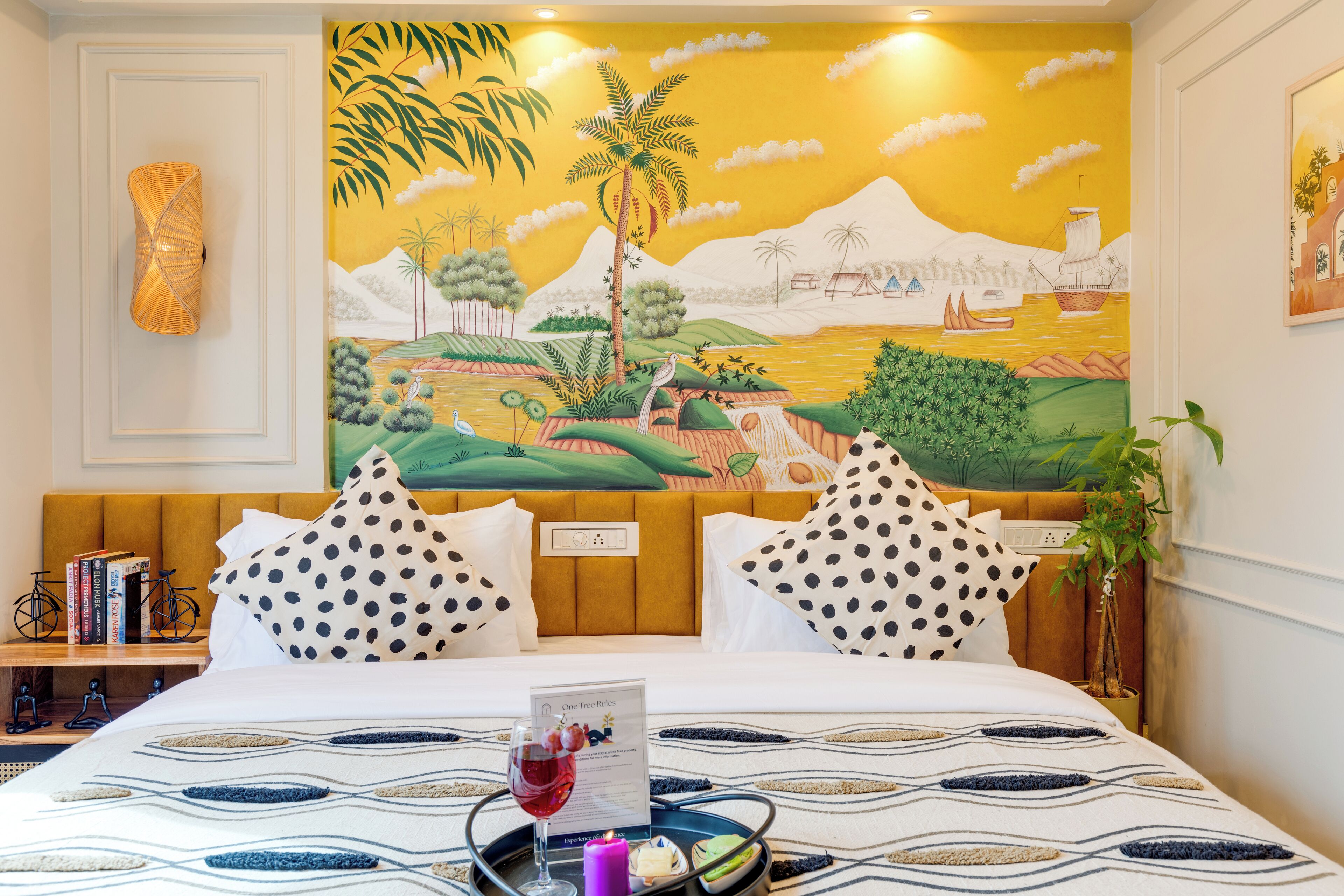 A hotel room with a bed, polka dot pillows, a nightstand with books, a wall mural, and a tray with a drink and candle.
