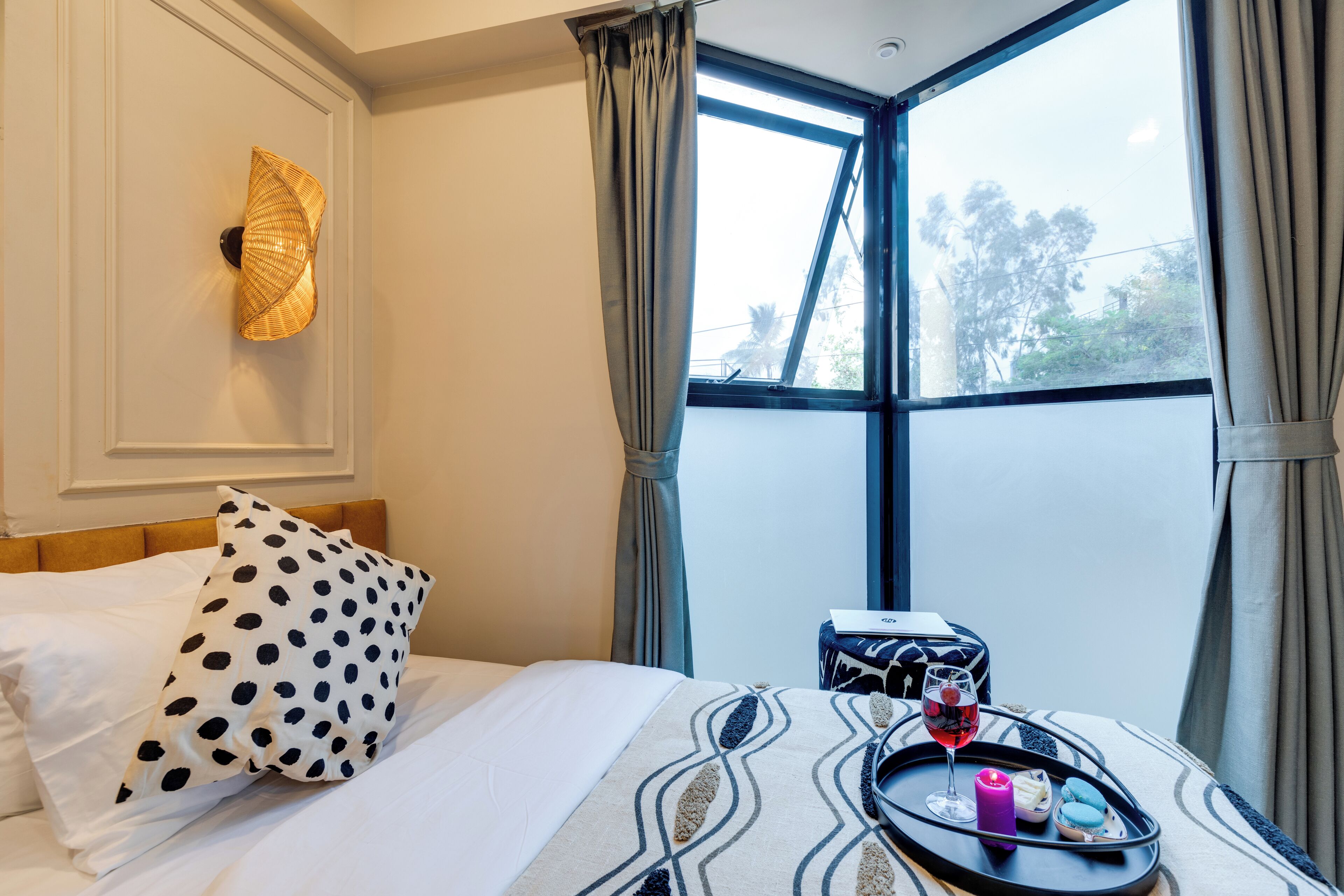 A hotel room with a bed, a bedside table with a tray of snacks, a decorative wall light, and a large window with curtains.