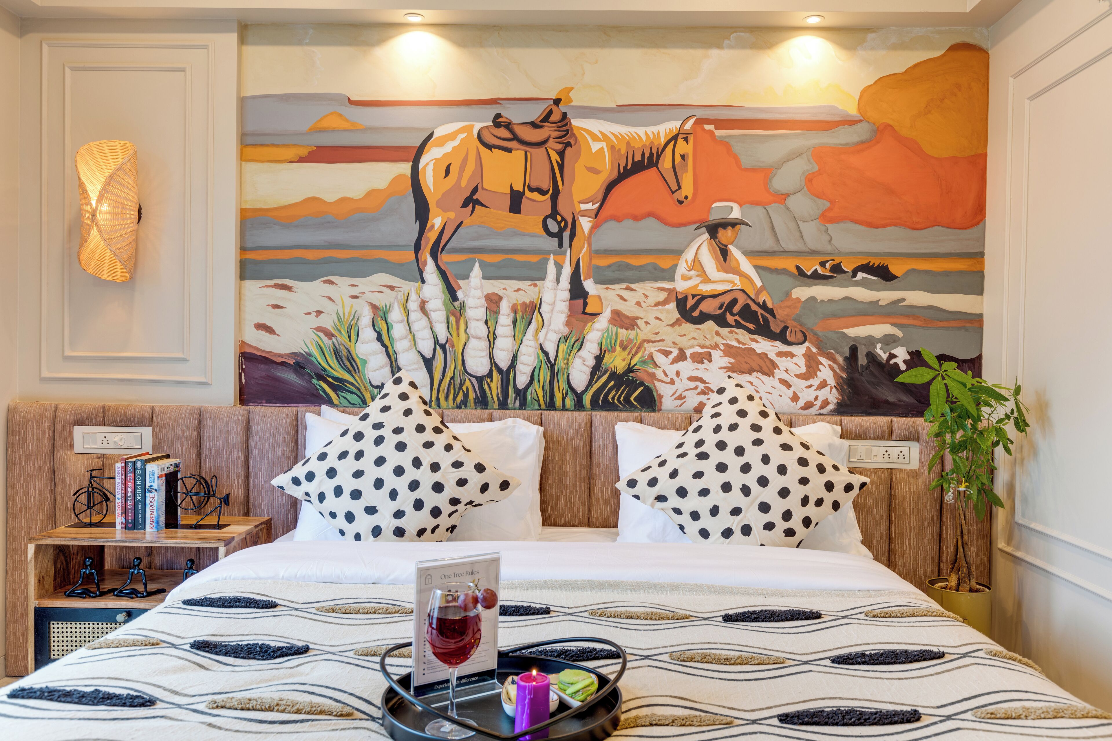 Ein Hotelzimmer mit einem großen Gemälde eines Reiters auf der Wand, einem Bett mit einem gemusterten Überwurf und einem Nachttisch mit Büchern und einer Kerze.