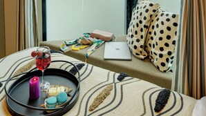Una habitación de hotel con un ventanal grande, una cama con un almohadón estampado de lunares, una computadora portátil y una bandeja con una vela, una copa de vino tinto y macarons.