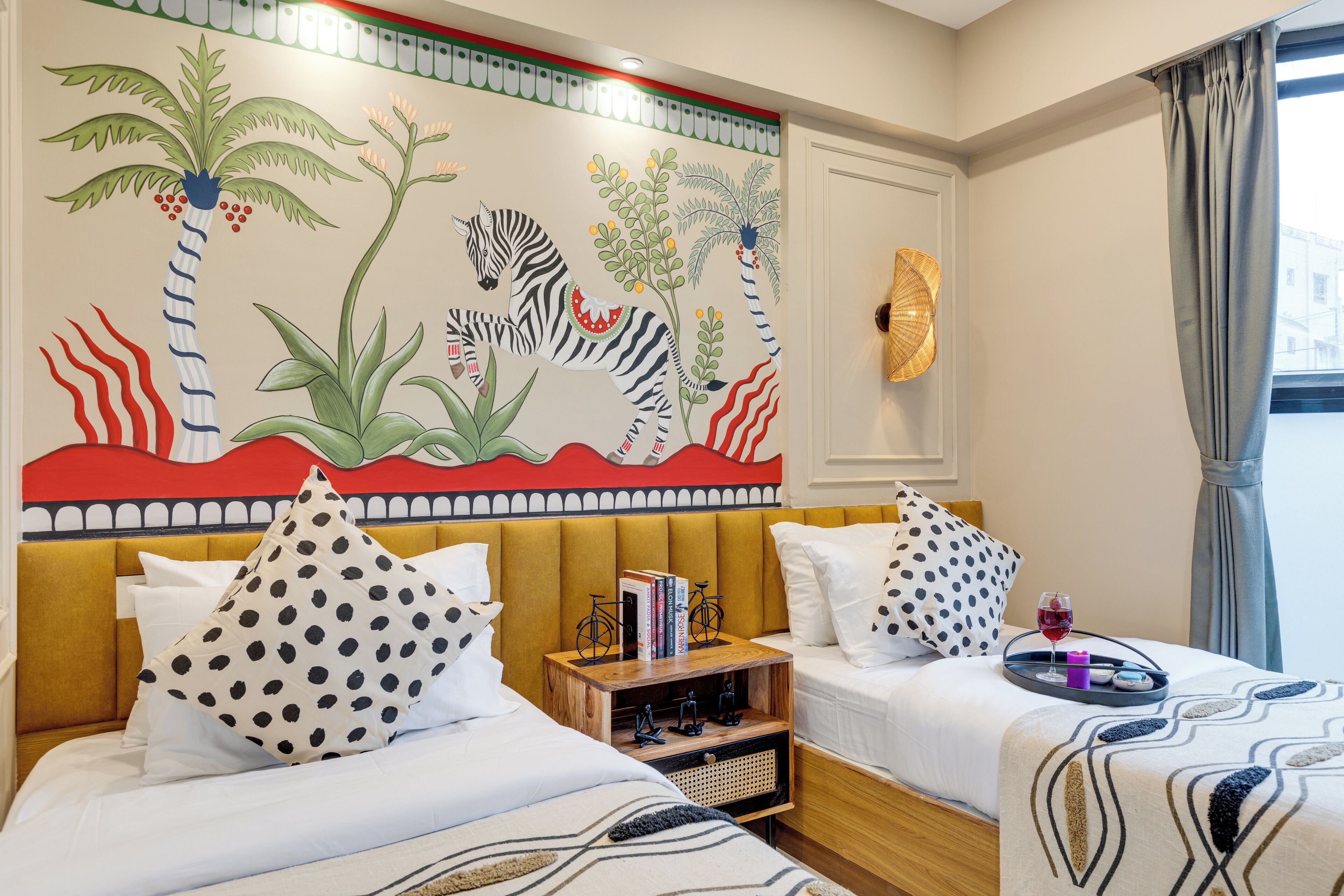 A hotel room with two beds, a mural on the wall, and a small wooden side table with books and a tray.