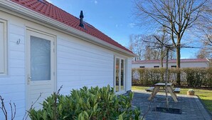 A white building with a red roof, a door, and a window. There is a patio area with a table and chairs, and a tree in the background. - Chalet in Friesland Near Large Lakes (Sint Nicolaasga)