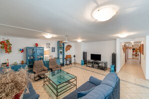 Un salon comprenant un canapé, un fauteuil, une table basse en verre et un téléviseur. On y trouve également une bibliothèque et une porte qui donne sur une autre pièce.