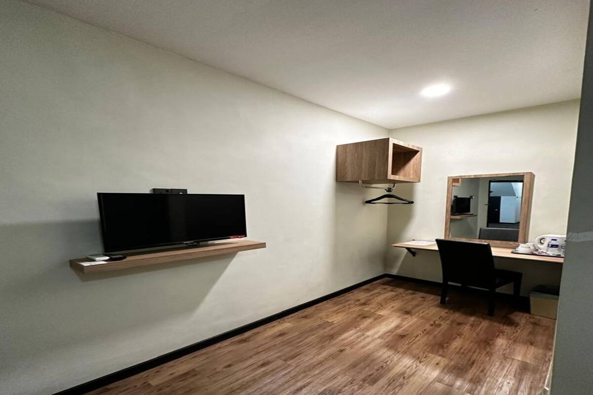 Une petite pièce moderne, dotée d’un téléviseur à écran plat posé sur une étagère en bois, d’un bureau avec une chaise, d’un miroir et d’une armoire en bois.