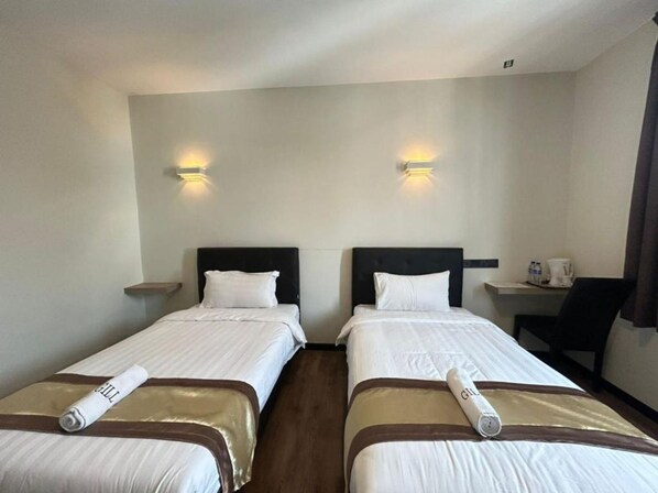 A hotel room with two single beds, each with a neatly rolled towel and a headboard. There are wall-mounted light fixtures above each bed and a small table with a chair to the right. - Gill Garden Boutique Hotel (Malacca City)
