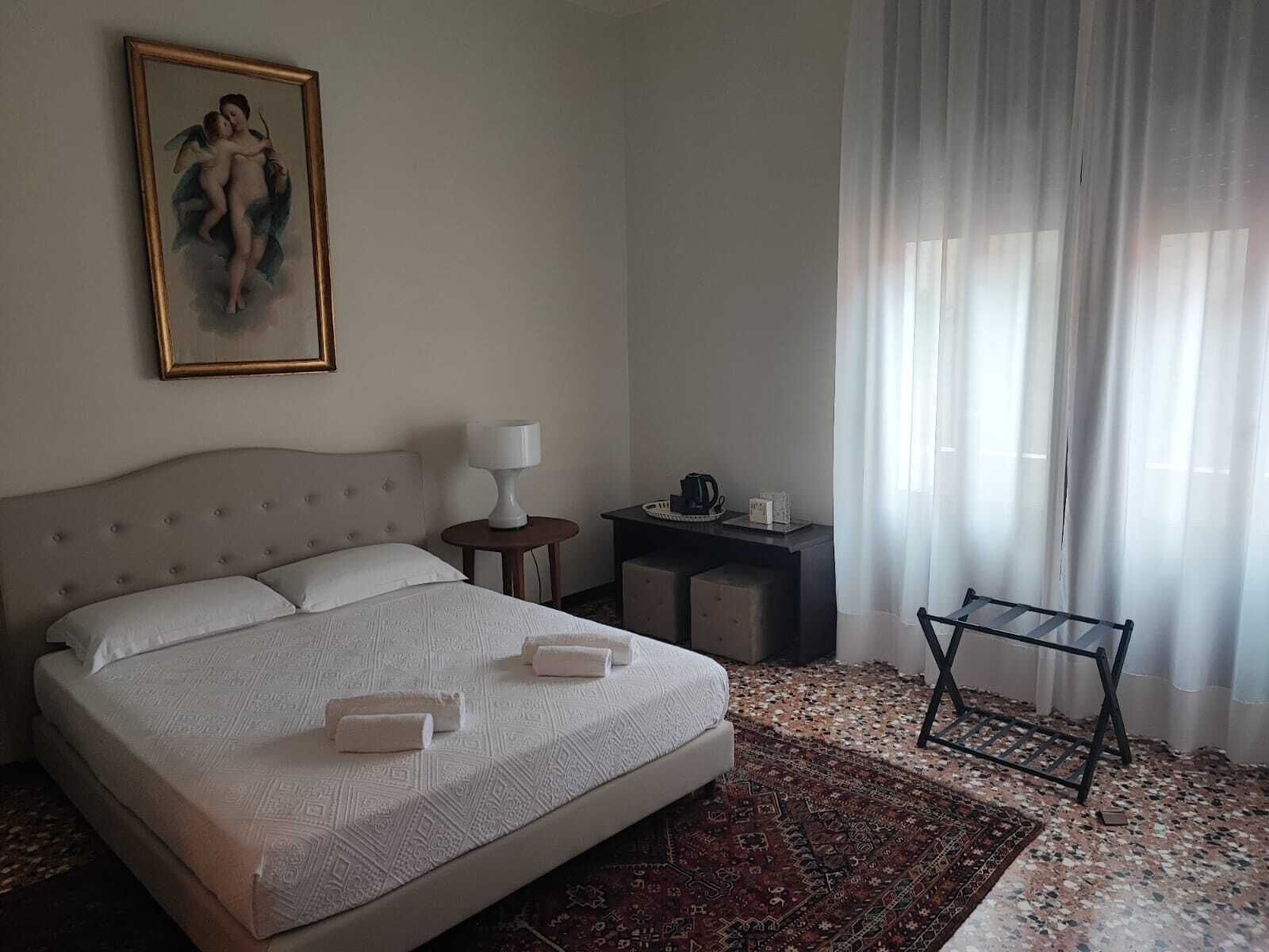 Habitación de hotel con una cama grande, una mesita de noche con lámpara, una pequeña mesa con cafetera y una ventana con cortinas translúcidas.