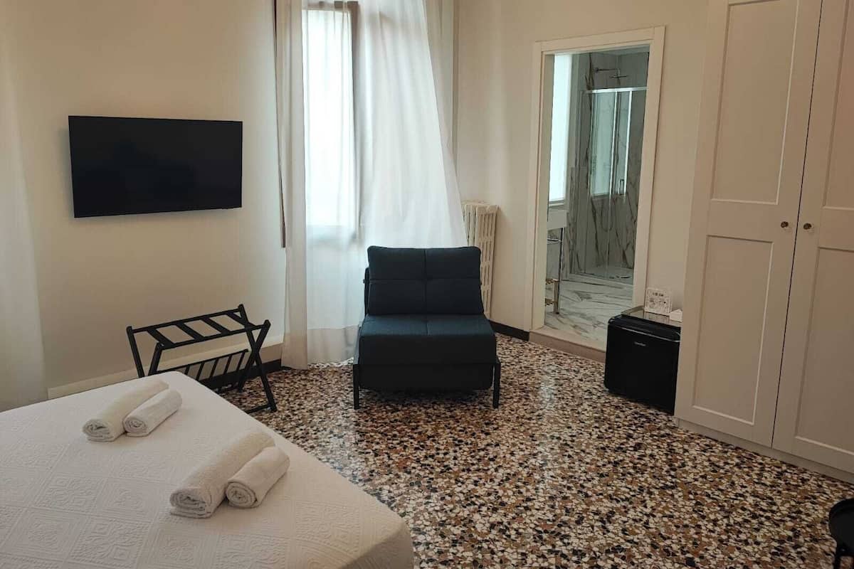 A hotel room with a bed, a chair, a TV, a door, and a view of a hallway.