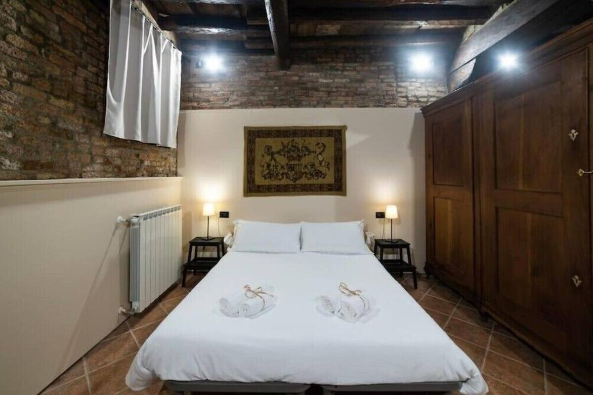 A hotel room with a large bed, two bedside tables with lamps, a radiator, a wooden wardrobe, and a brick wall with a decorative tapestry.