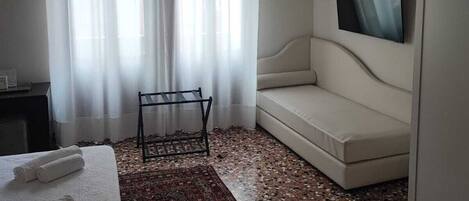 A hotel room with a bed, a sofa, a TV mounted on the wall, and a window with sheer curtains.