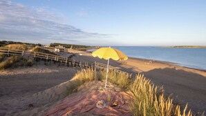 A beach with a wooden fence, a yellow umbrella, and a blanket with food and drinks.