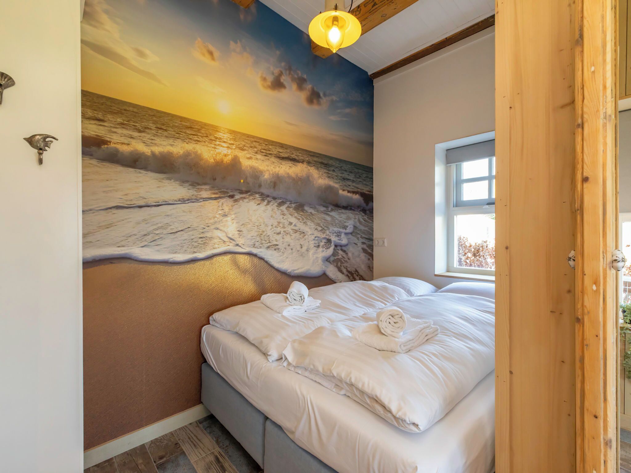 A bedroom with a mural of a beach scene, a bed with white linens, and a window with a view of the outdoors.