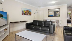 Un salon avec un canapé en cuir noir, une table basse blanche, un téléviseur posé sur un meuble, un tableau et un miroir au mur.