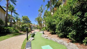 Un chemin bordé d’herbe artificielle mène à un bâtiment, lequel est entouré d’une végétation luxuriante et de palmiers.