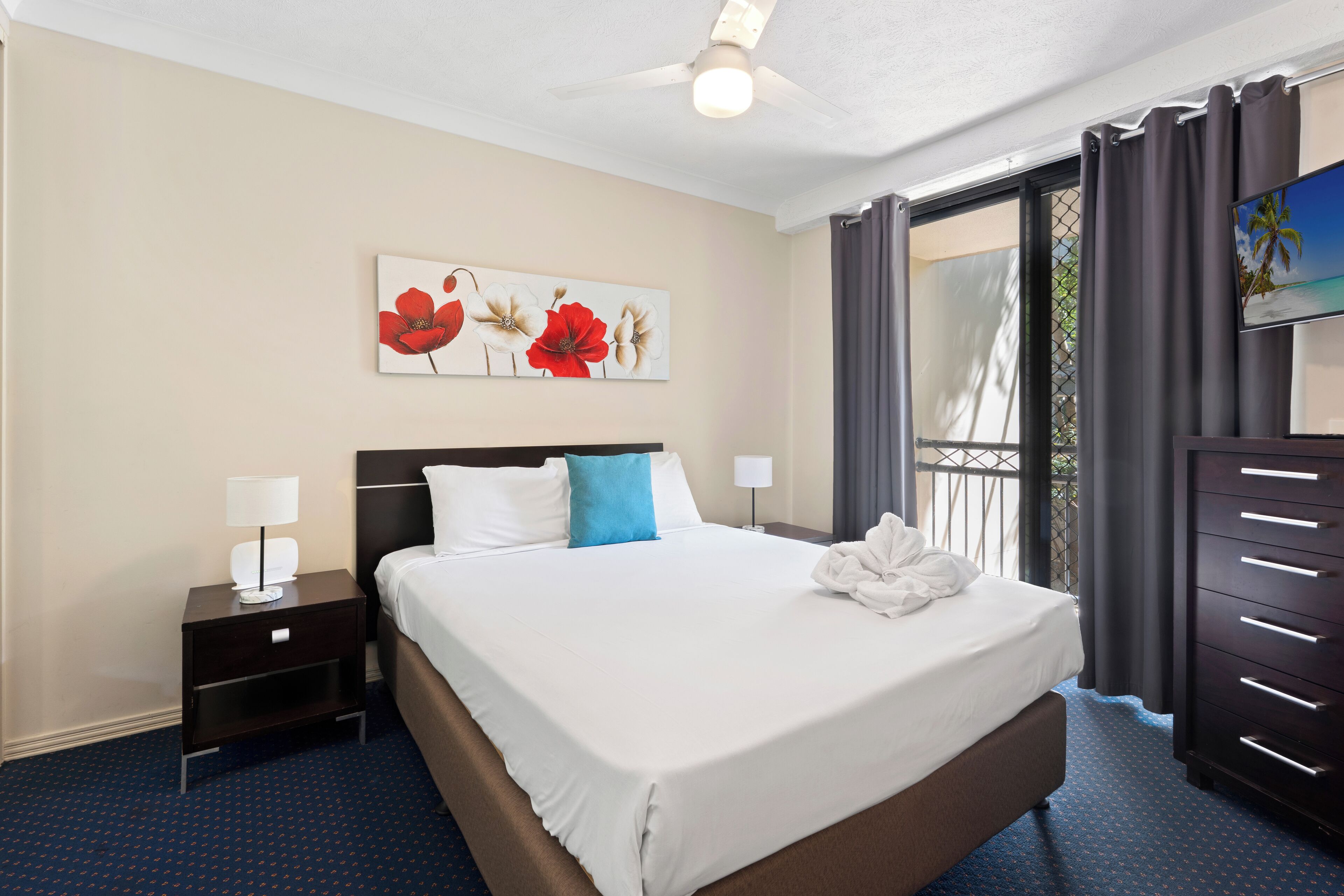 A hotel room with a large bed, a nightstand with a lamp, a painting of flowers on the wall, and a TV on the dresser.