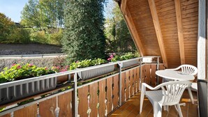 Un balcon en bois avec une table et des chaises blanches, entouré de plantes en pot et offrant une vue sur les arbres.