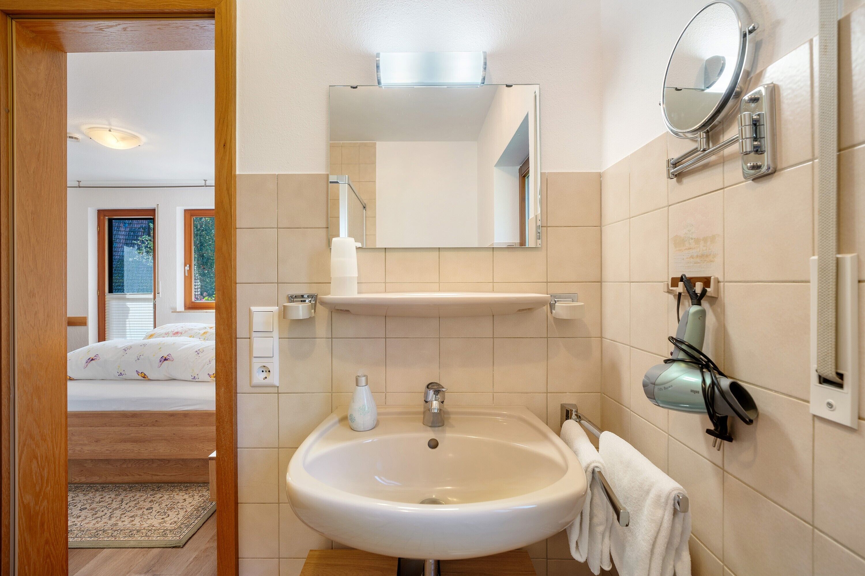 Baño con lavamanos, espejo, toallero y zona de ducha visible a través de una puerta abierta.