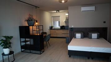 A modern hotel room with a bed, a desk with a chair, a potted plant, and a kitchen area in the background.